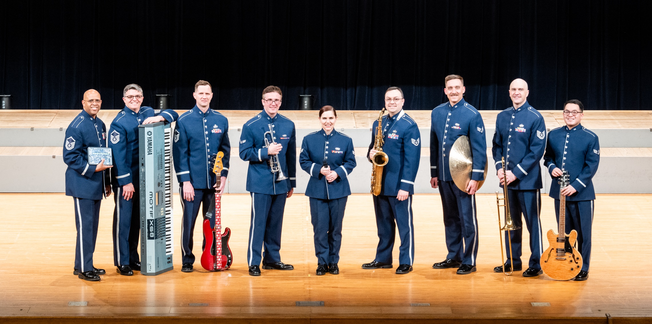 Nine members of the Final Approach rock ensemble stand on a wooden stage wearing their dark blue tunic uniforms with silver buttons and stripes, holding a variety of rock instruments including guitars and pianos.